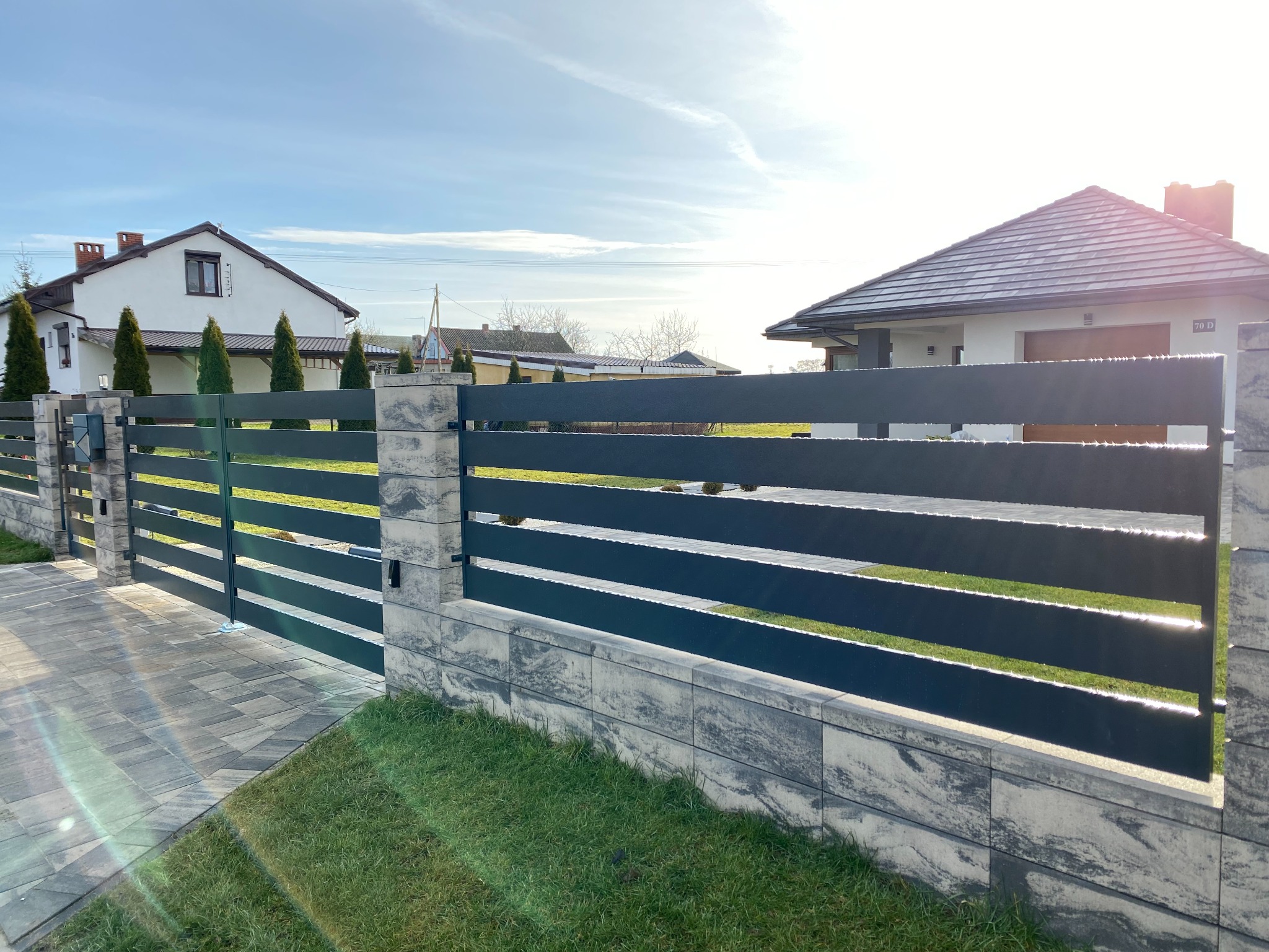 Nowoczesne, poziome ogrodzenie panelowe w kolorze antracytowym, osadzone na murku z szarej kostki imitującej kamień, z bramą wjazdową, na tle zadbanego trawnika i dwóch domów jednorodzinnych...