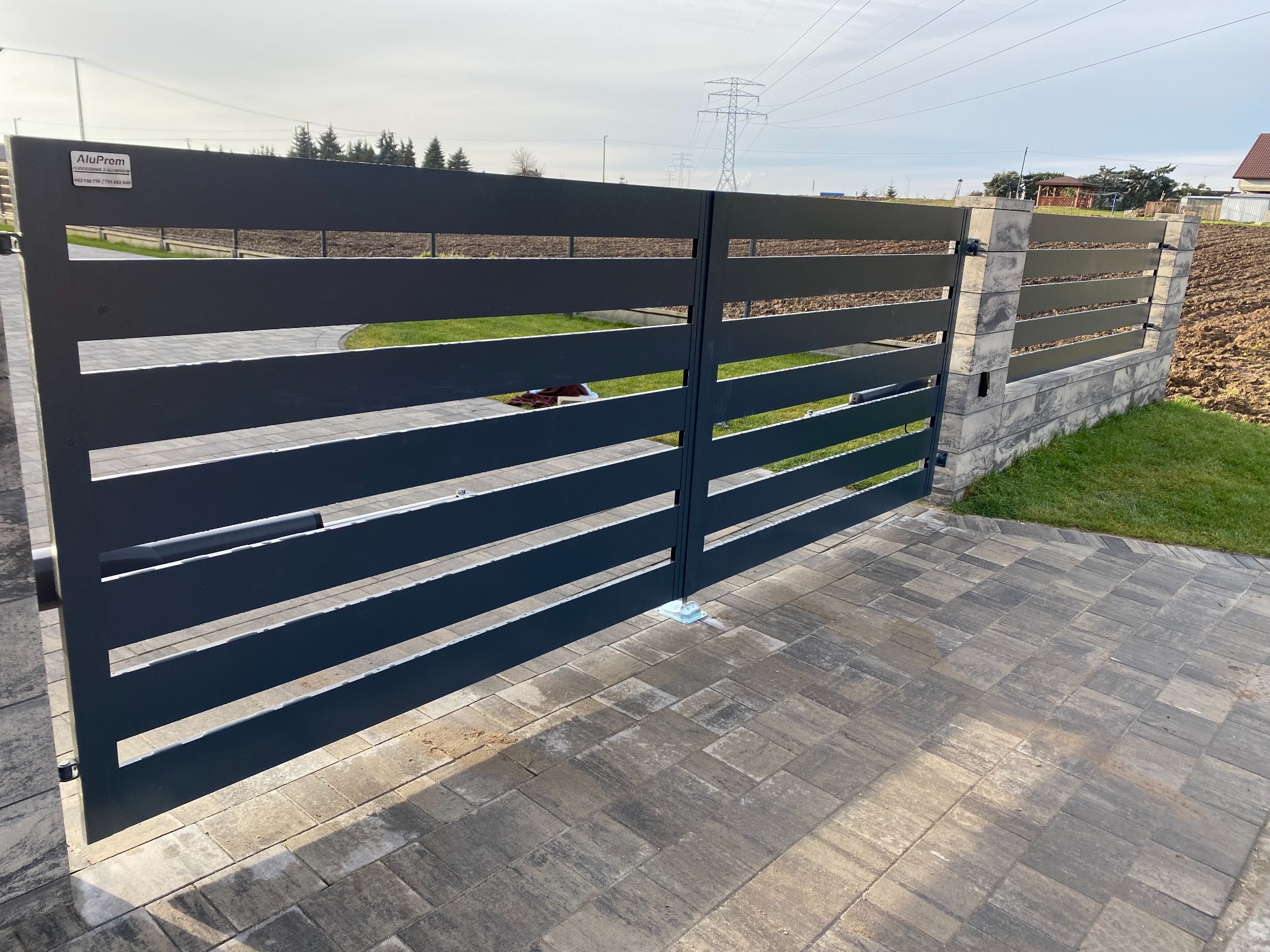 Nowoczesna, pozioma brama wjazdowa z szarych, aluminiowych profili, zamontowana na betonowych słupkach z kostki, widok z zewnątrz na podjeździe z kostki brukowej, w tle trawnik i dom.