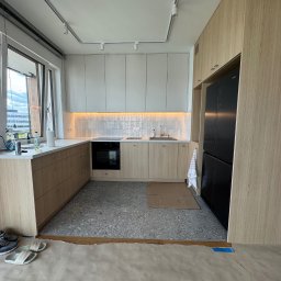Helilab Sp. z o.o. - Jasna kuchnia w trakcie montażu: drewniane szafki, szare płytki podłogowe, zabezpieczona podłoga. Widoczne buty i klapki. Minimalistyczny design, nowoczesne oświetlenie.