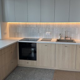 Helilab Sp. z o.o. - Jasna kuchnia z drewnianymi szafkami, białym blatem i podświetlanymi płytkami. Nowoczesny design, widok z okna, sprzęt AGD w zabudowie, armatura kuchenna.