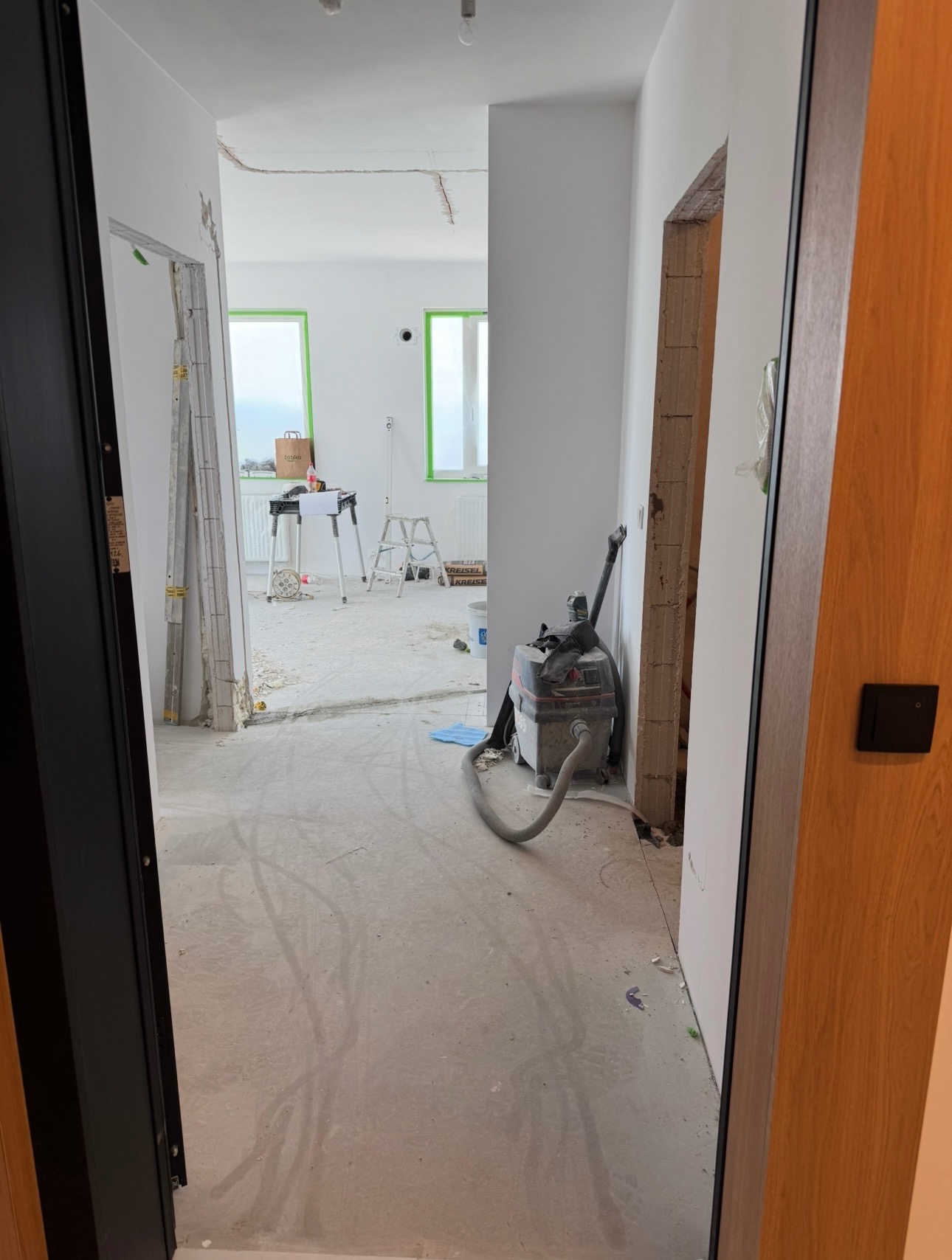 Wnętrze w trakcie remontu: odnowione ściany, zabezpieczone okna folią, widoczny odkurzacz przemysłowy i narzędzia, surowy beton na podłodze, otwarte przejście.