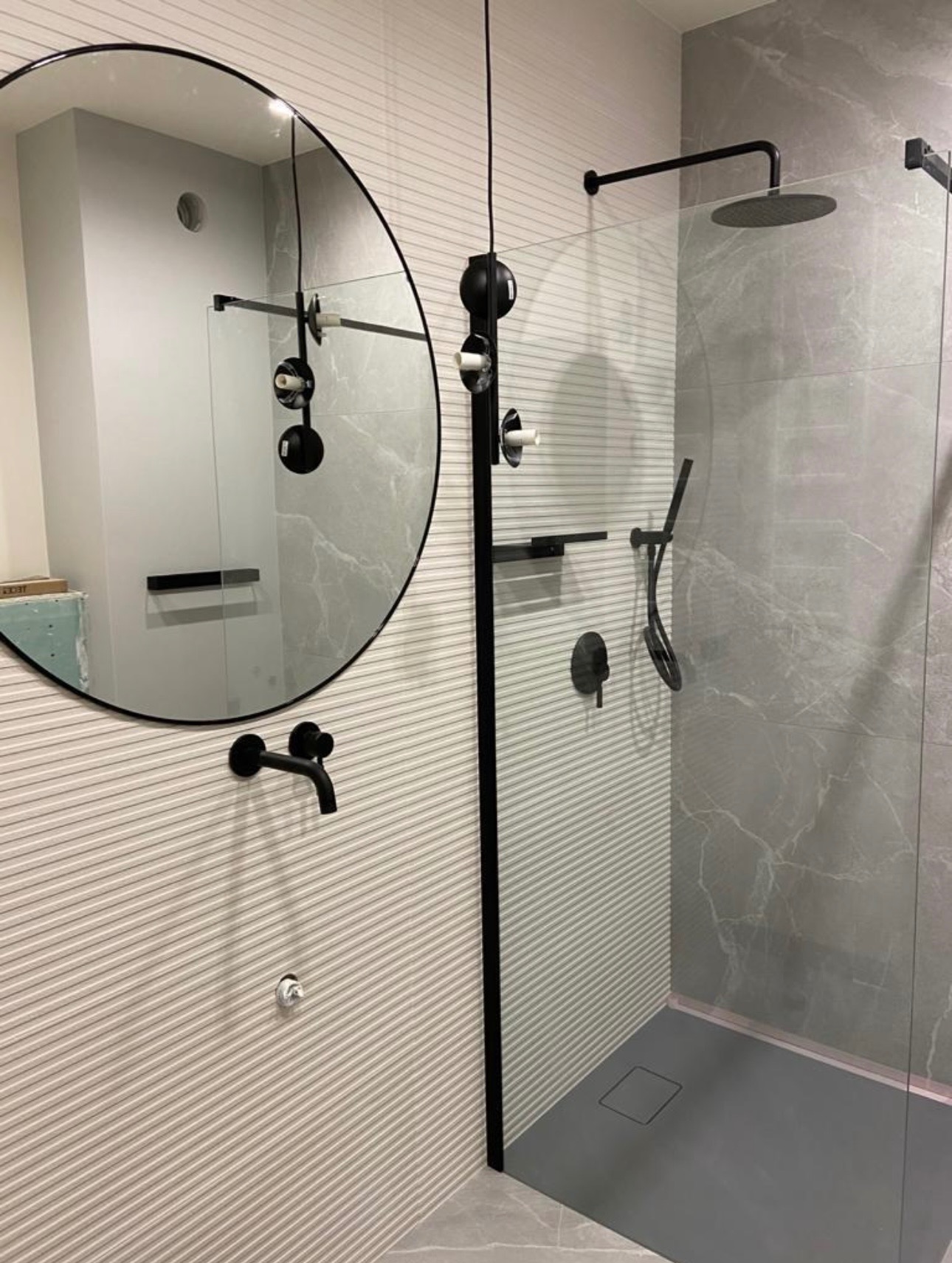 Nowoczesna łazienka w Warszawie z kabiną prysznicową typu walk-in, okrągłym lustrem i czarną armaturą. Wykończenie ścian płytkami w odcieniach szarości.