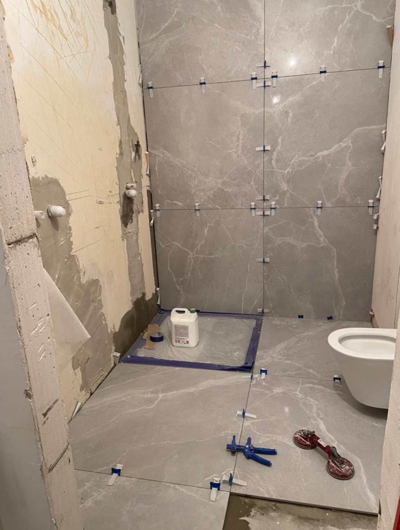 Układanie szarych płytek imitujących marmur w łazience. Widoczne narzędzia glazurnicze i poziomowanie płytek. Częściowo wykończona ściana i podłoga.