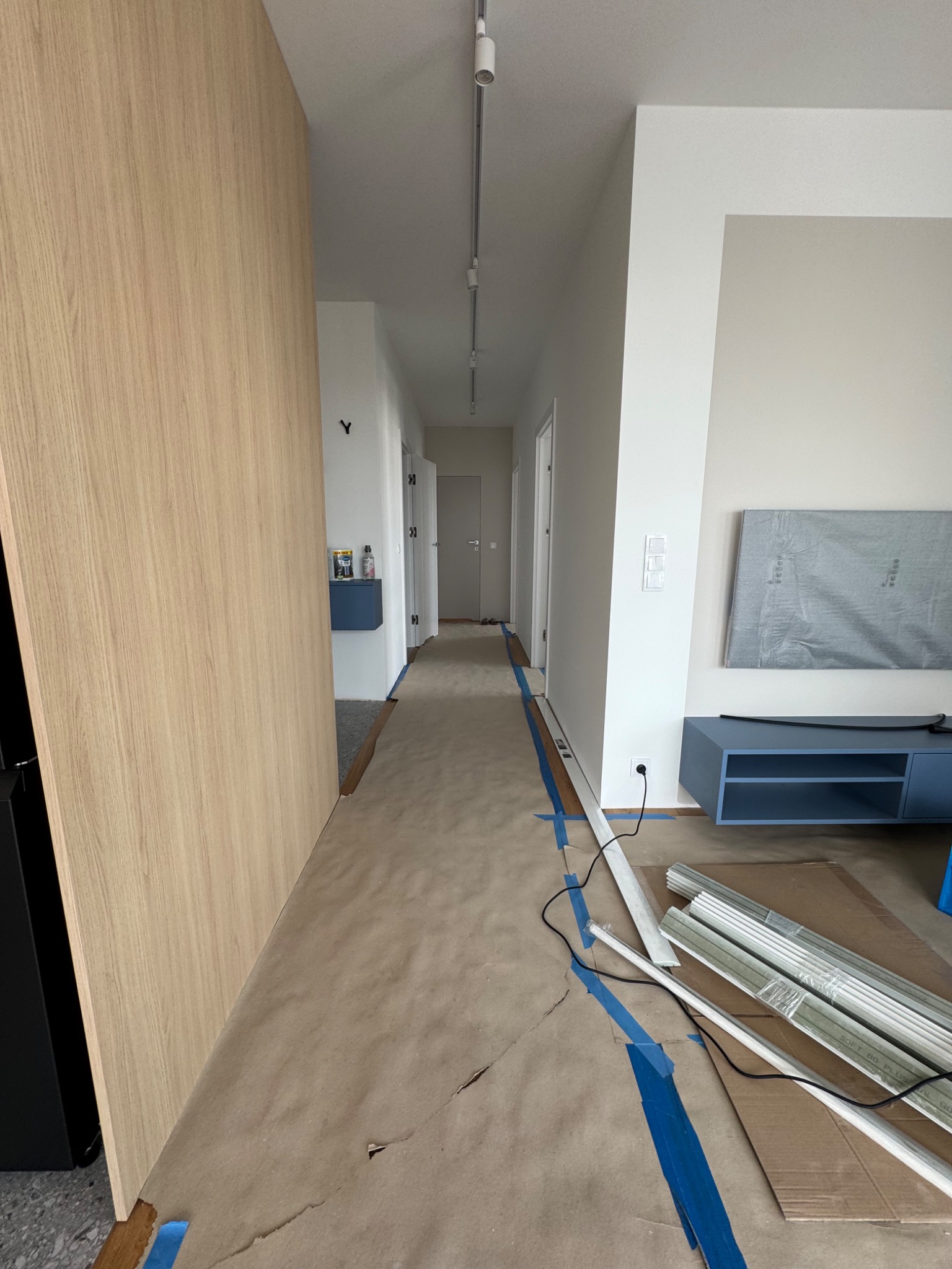 Przygotowanie do montażu listew przypodłogowych w jasnym wnętrzu. Podłoga zabezpieczona kartonem i niebieską taśmą malarską. Widoczne listwy i narzędzia.