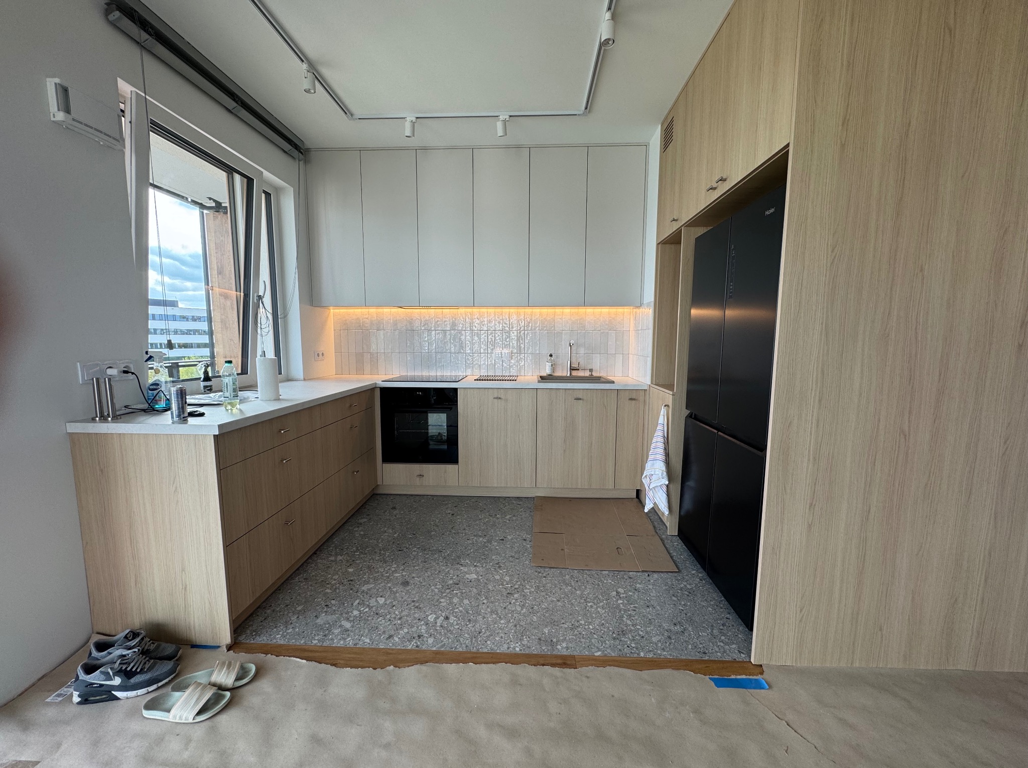 Jasna kuchnia w trakcie montażu: drewniane szafki, szare płytki podłogowe, zabezpieczona podłoga. Widoczne buty i klapki. Minimalistyczny design, nowoczesne oświetlenie.