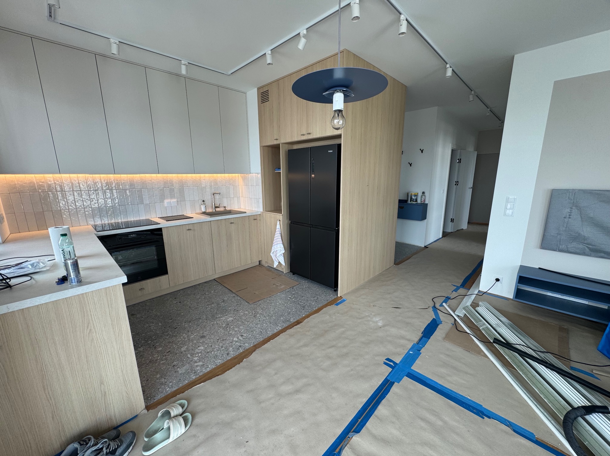 Jasna kuchnia z drewnianymi szafkami i szarą podłogą w trakcie remontu. Widoczne zabezpieczenia podłogi i listwy przypodłogowe. Nowoczesne oświetlenie sufitowe.