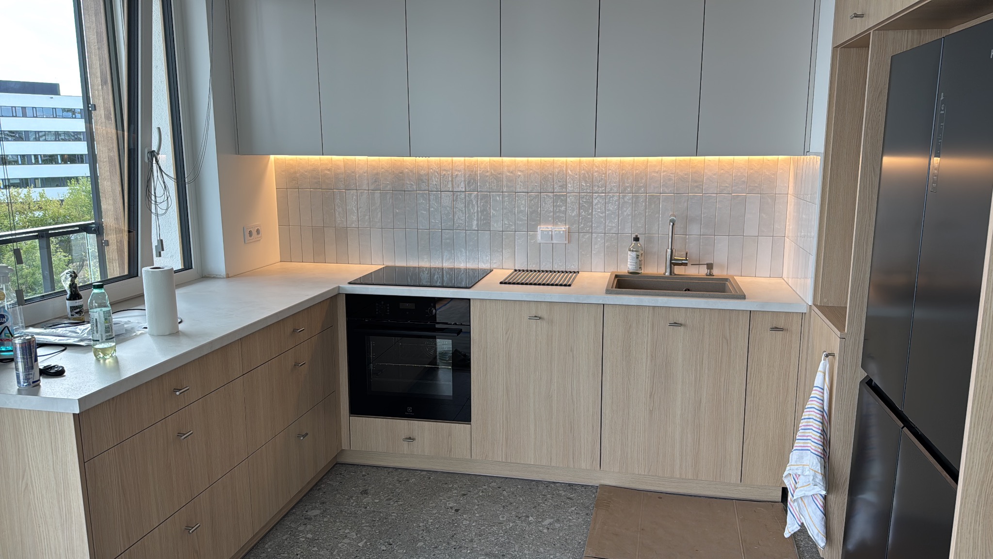 Jasna kuchnia z drewnianymi szafkami, białym blatem i podświetlanymi płytkami. Nowoczesny design, widok z okna, sprzęt AGD w zabudowie, armatura kuchenna.