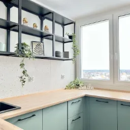 Jasna kuchnia z miętowo-zielonymi szafkami, drewnianym blatem, białymi płytkami imitującymi lastryko, czarnymi otwartymi półkami i roślinami doniczkowymi, widok na miasto za oknem.