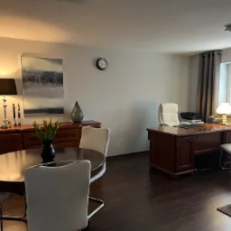 Jasne biuro z ciemnym drewnianym biurkiem, okrągłym stołem z tulipanami i białymi krzesłami na ciemnej podłodze.