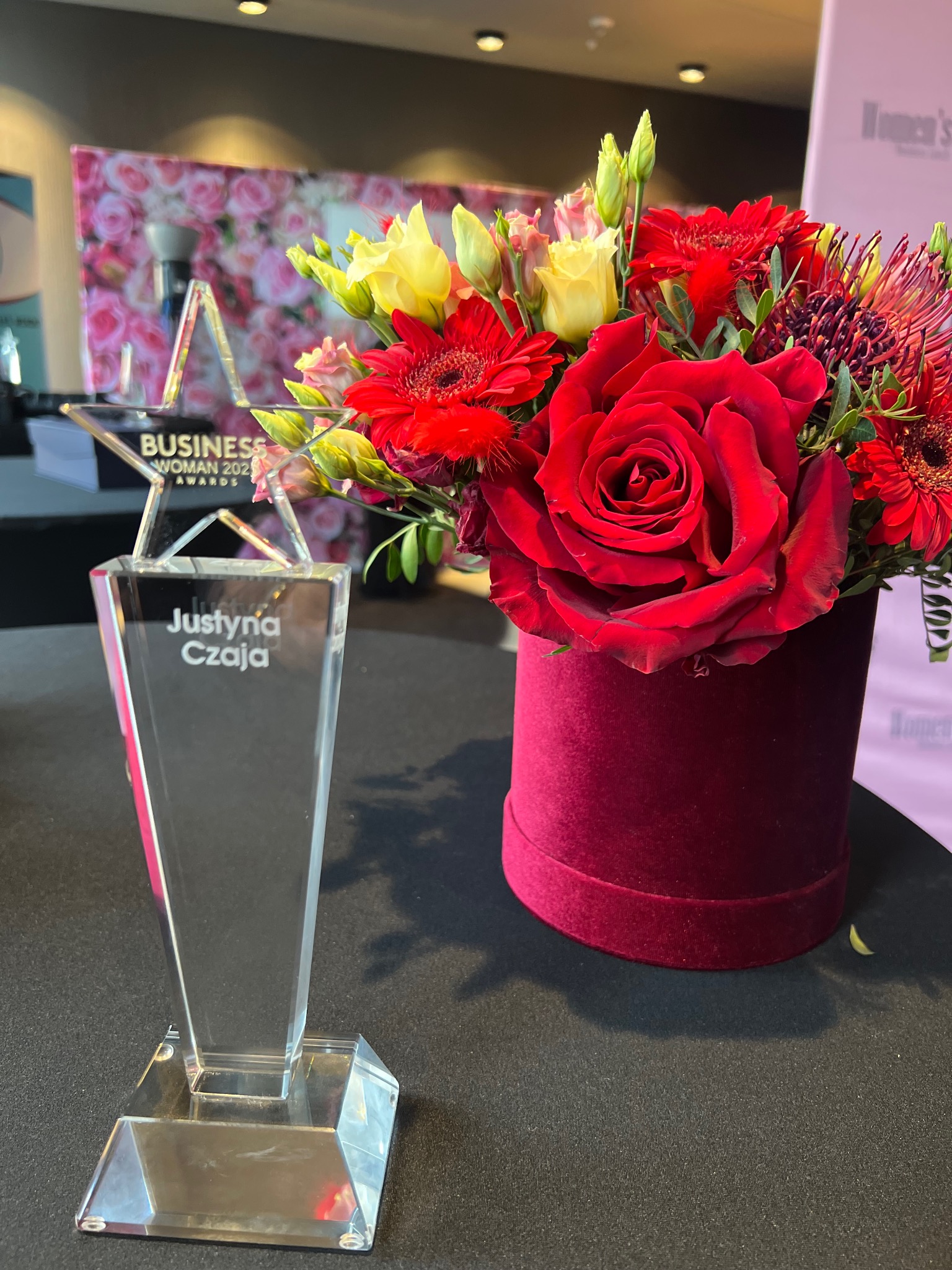 Kryształowa statuetka 'Business Woman 2023 Awards' z imieniem Justyna Czaja, obok bukiet czerwonych róż w bordowym, welurowym pudełku na czarnym stole.