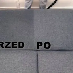 Sofa z wyraźnym podziałem na część wyczyszczoną i zabrudzoną, z napisem 'PRZED' i 'PO', widoczny fragment węża odkurzacza piorącego.