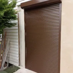 Zamknięta, brązowa roleta zewnętrzna zamontowana na ścianie budynku z widoczną elewacją i fragmentem roślinności po lewej stronie.