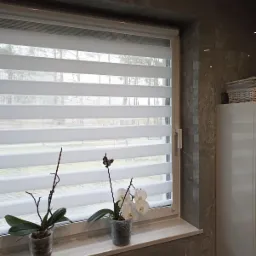 Okno z białymi roletami zebra i dwoma doniczkami z orchideami na marmurowym parapecie w łazience wykończonej płytkami imitującymi kamień.