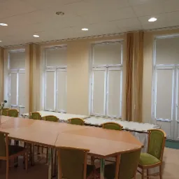 Sala konferencyjna z długim stołem i krzesłami, okna przesłonięte jasnymi roletami zwijanymi do góry.