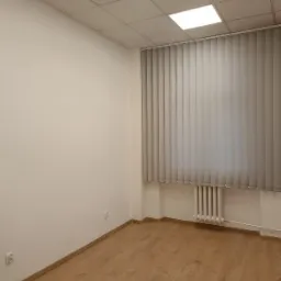 Wnętrze biurowe z pionowymi lamelami okiennymi w kolorze szarym, zamontowanymi na całą wysokość okna, nad grzejnikiem.