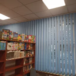 Błękitne pionowe lamele w oknie biblioteki, z widocznym regałem z książkami i grami planszowymi w tle.