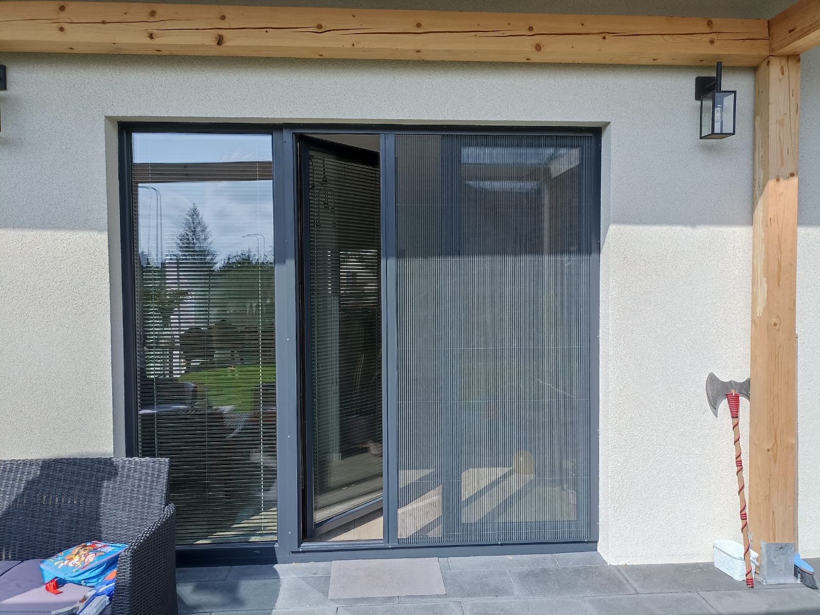 Zamknięta przesuwna moskitiera plisowana w kolorze szarym, zamontowana w przeszklonym otworze drzwiowym, widok z zewnątrz tarasu, z drewnianą belką stropową i lampą ścienną.