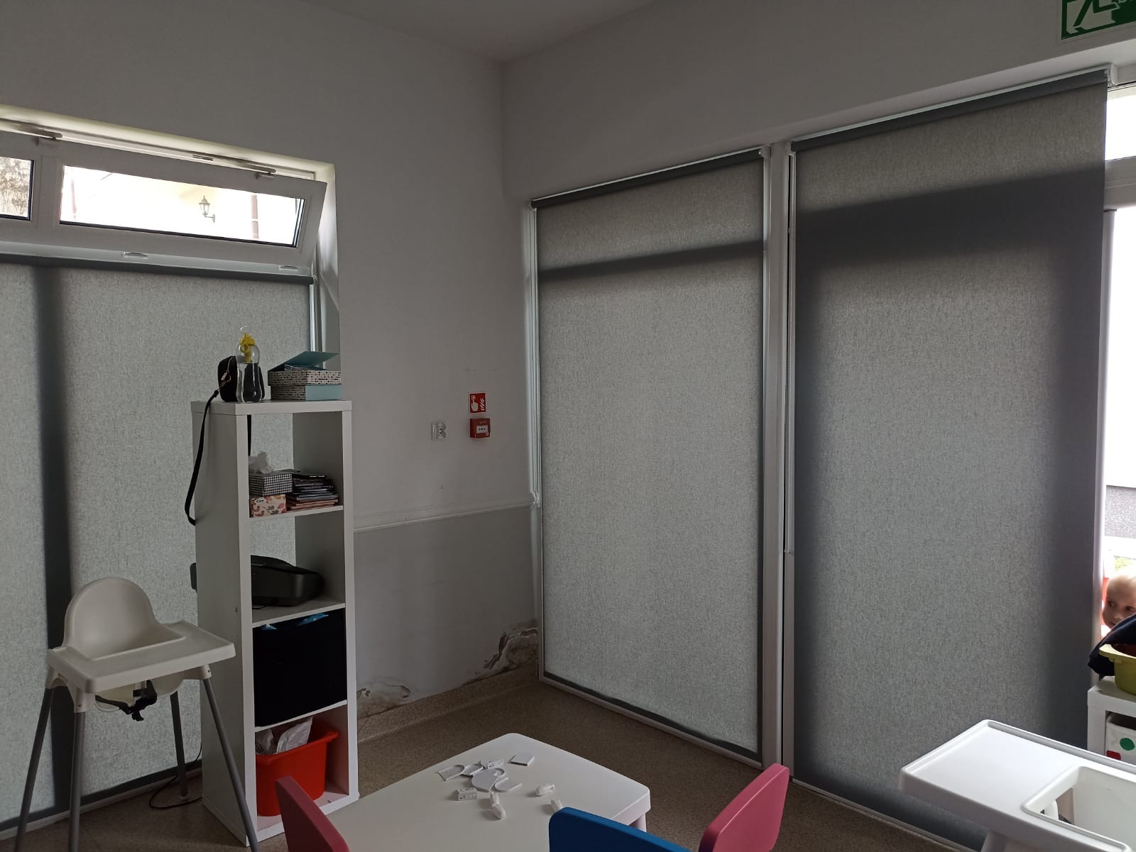 Szare rolety wewnętrzne w pokoju dziecięcym, widoczne meble dziecięce i zabawki, ściany z widocznymi śladami użytkowania.