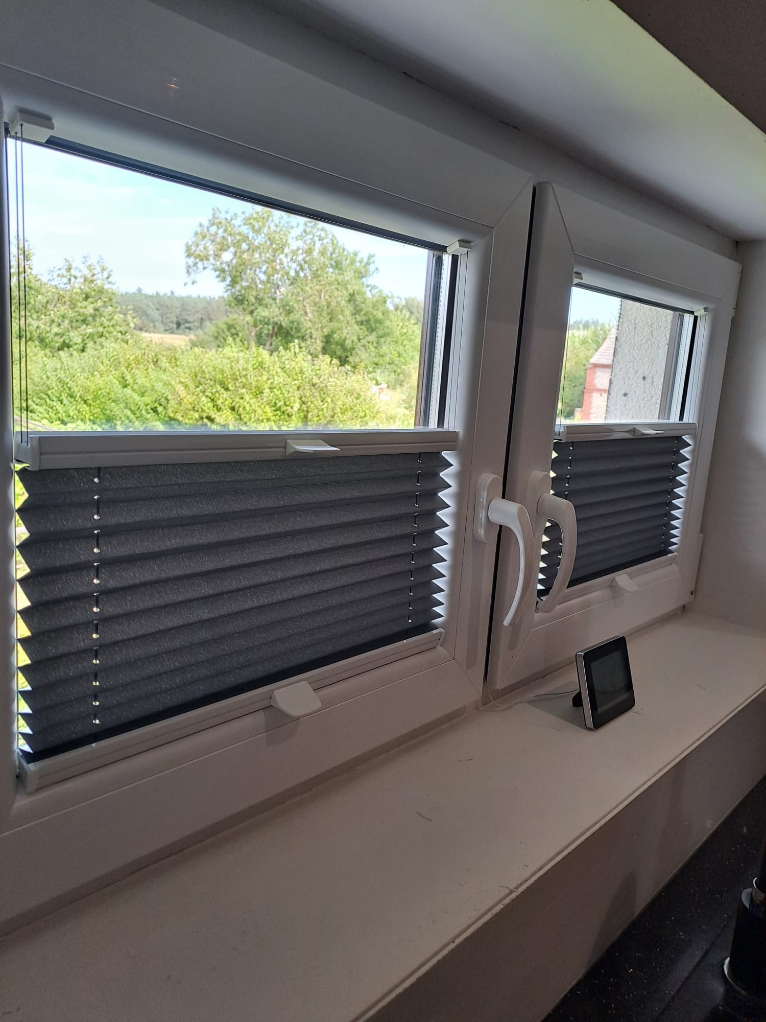 Szare, plisowane osłony okienne zamontowane w oknach PCV, widok na zieleń za oknem.