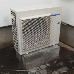 Zewnętrzna jednostka pompy ciepła Panasonic R32, zamontowana na betonowym cokole przy ścianie budynku z oknem i skrzynką elektryczną. Widoczna kostka brukowa na podłożu.