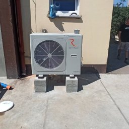 eco instalacje Paweł Engelke - Zewnętrzna jednostka pompy ciepła Rotenso Aquami zamontowana na betonowych bloczkach przy ścianie budynku z małym oknem, widoczne rury spustowe i fragment chodnika.