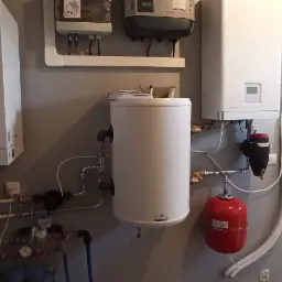 Węzeł cieplny z dwoma piecami, zbiornikiem buforowym i czerwonym naczyniem przeponowym w pomieszczeniu technicznym, rury prowadzone przy suficie.