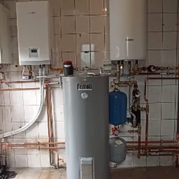 Kotłownia z trzema piecami różnych marek (Kaisai, Ferroli, inny) i rozbudowaną instalacją hydrauliczną z miedzianych rur na tle wykafelkowanej ściany.