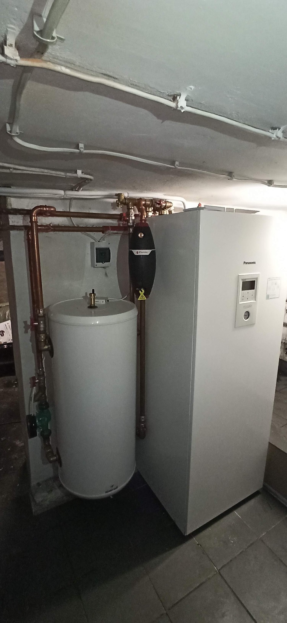 Zainstalowana pompa ciepła Panasonic Aquarea z zbiornikiem buforowym i osprzętem hydraulicznym, widoczne miedziane rury instalacji w piwnicy.