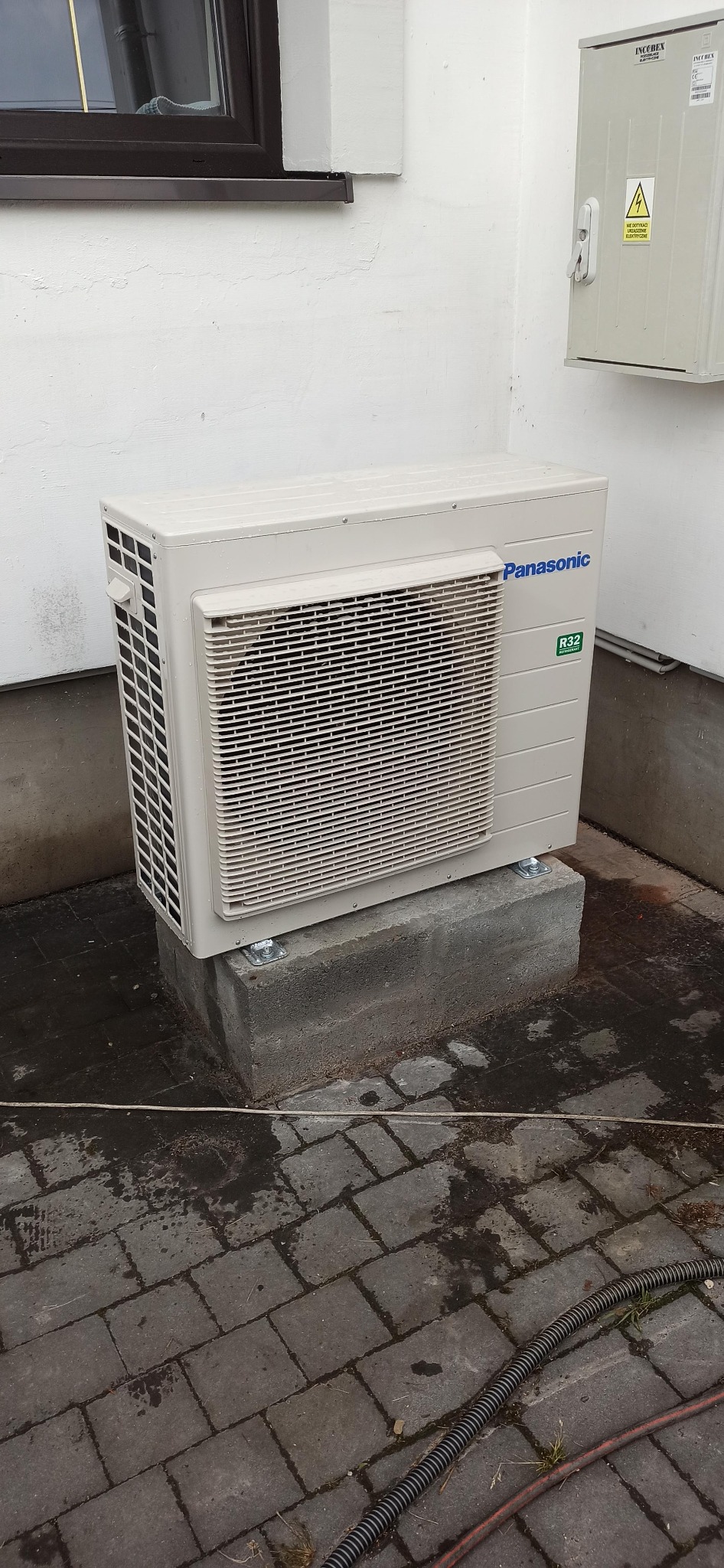 Zewnętrzna jednostka pompy ciepła Panasonic R32, zamontowana na betonowym cokole przy ścianie budynku z oknem i skrzynką elektryczną. Widoczna kostka brukowa na podłożu.