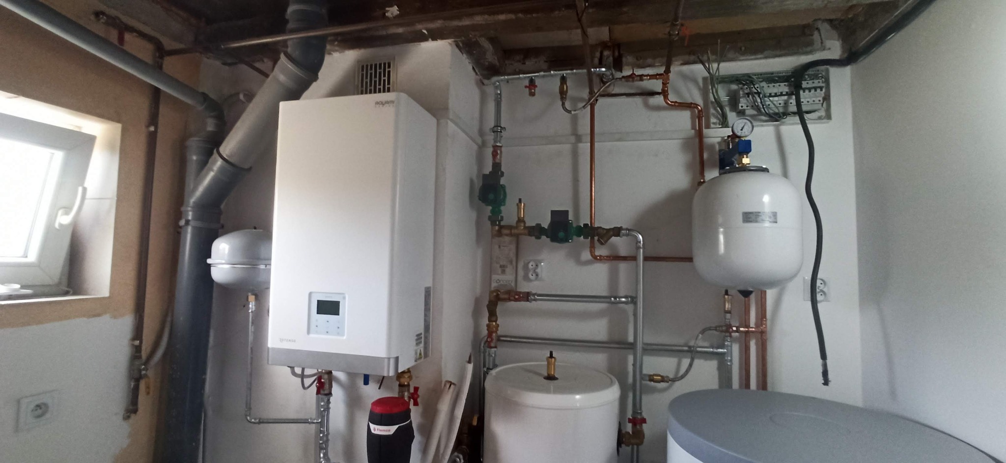 Biały piec gazowy zamontowany w kotłowni, widoczne rury miedziane, grzejniki, zawory, zbiornik wyrównawczy i instalacja elektryczna.