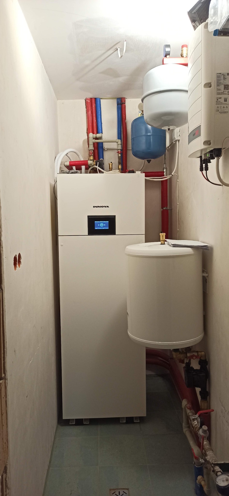Biała pompa ciepła marki Innova z podłączonymi rurami w kolorze czerwonym i niebieskim, widoczny zbiornik wyrównawczy i zasobnik ciepłej wody, instalacja w pomieszczeniu technicznym o surowych...