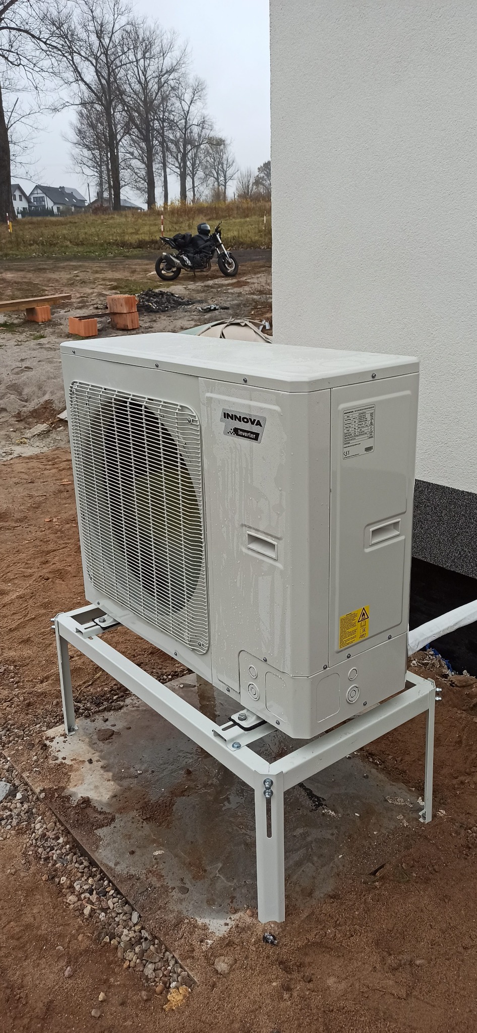 Zewnętrzna jednostka pompy ciepła Innova Inverter, zamontowana na metalowym stelażu, z motocyklem w tle i widocznymi elementami instalacji.