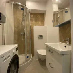 Nowoczesna łazienka z kabiną prysznicową, umywalką z szafką w kolorze białym, toaletą i pralką Amica, wykończona płytkami w odcieniach bieli i drewna ułożonymi we wzór jodełki.