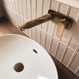 Biała, okrągła umywalka z złotą armaturą Hansgrohe na tle beżowych, pionowych płytek ułożonych w cegiełkę, z drewnianym blatem pod umywalką.