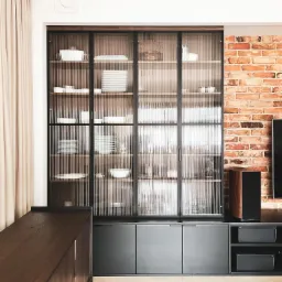 Nowoczesna aranżacja salonu z przeszkloną witryną w czarnej ramie, wypełnioną białą porcelaną, na tle ściany z czerwonej cegły i drewnianej komody.