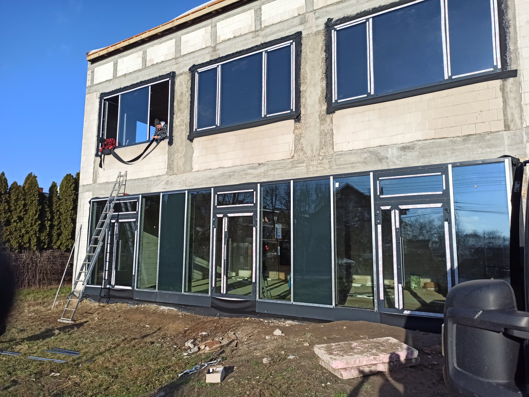 Budynek w trakcie montażu okien i drzwi, widoczny pracownik uszczelniający okno na piętrze, drabina oparta o fasadę.