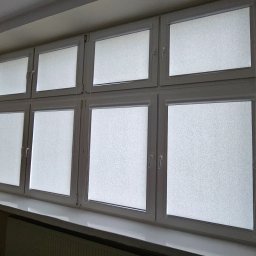Firma AMS OKNA - Białe okna PCV z roletami wewnętrznymi w budynku. Widoczne parapety i fragment ściany z tekstem. Ujęcie z dołu, podkreślające regularny układ okien.