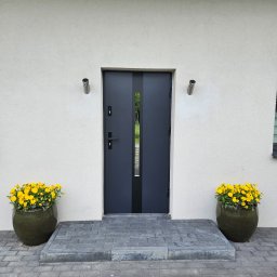 Firma AMS OKNA - Eleganckie, grafitowe drzwi wejściowe z przeszkleniem, flankowane donicami z żółtymi kwiatami. Nowoczesne oświetlenie elewacyjne. Kostka brukowa prowadzi do wejścia.