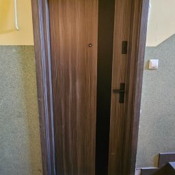 Firma AMS OKNA - Brązowe drzwi wejściowe z czarnym pasem pośrodku, zamontowane w bloku mieszkalnym. Widoczne schody i fragment ściany z przełącznikiem.