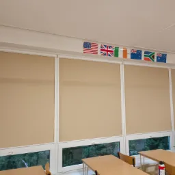 Beżowe rolety wewnętrzne w sali lekcyjnej w Radomsku, z widocznymi flagami różnych państw nad oknami.