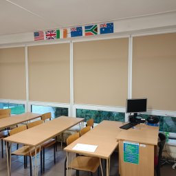 Firma AMS - Wnętrze sali lekcyjnej z beżowymi roletami zwiniętymi do połowy, dekorowanymi flagami różnych państw i zasłonami z nadrukowanymi napisami.