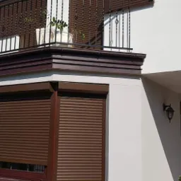 Brązowe rolety zewnętrzne zamontowane na oknach domu z balkonem, widoczne rośliny w donicach na balkonie.