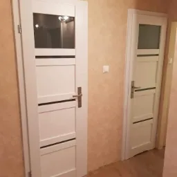 Dwa białe, nowoczesne skrzydła drzwiowe z przeszkleniami w górnej części, zamontowane w jasnym wnętrzu z beżowymi ścianami i brązową podłogą.