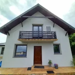 Dom jednorodzinny z balkonem, brązowymi drzwiami wejściowymi i szarą stolarką okienną, widoczna moskitiera w oknie na piętrze.