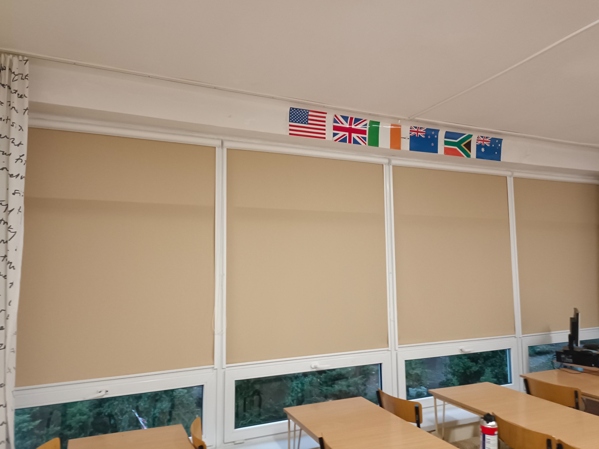 Beżowe rolety wewnętrzne w sali lekcyjnej w Radomsku, z widocznymi flagami różnych państw nad oknami.
