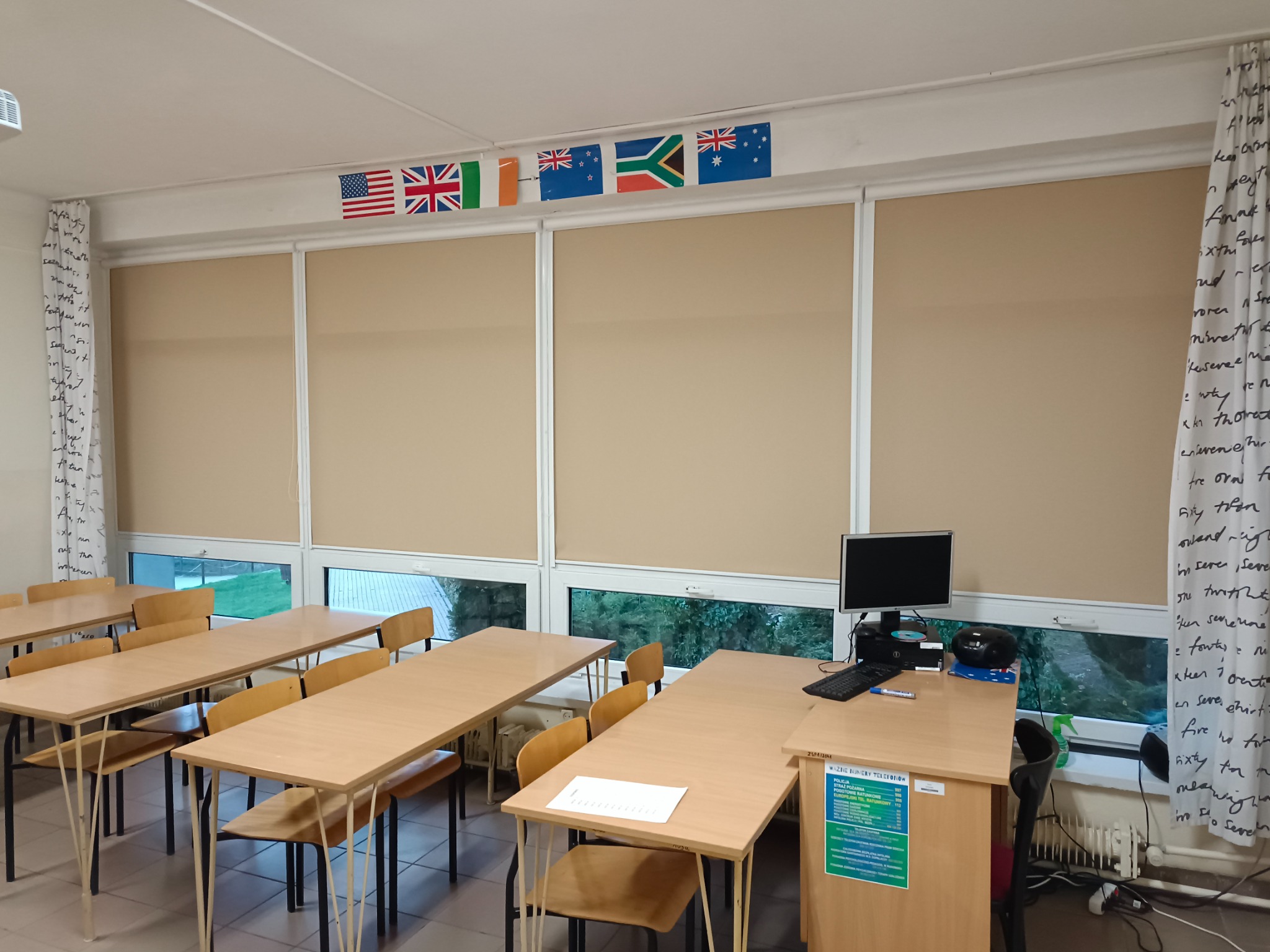 Wnętrze sali lekcyjnej z beżowymi roletami zwiniętymi do połowy, dekorowanymi flagami różnych państw i zasłonami z nadrukowanymi napisami.