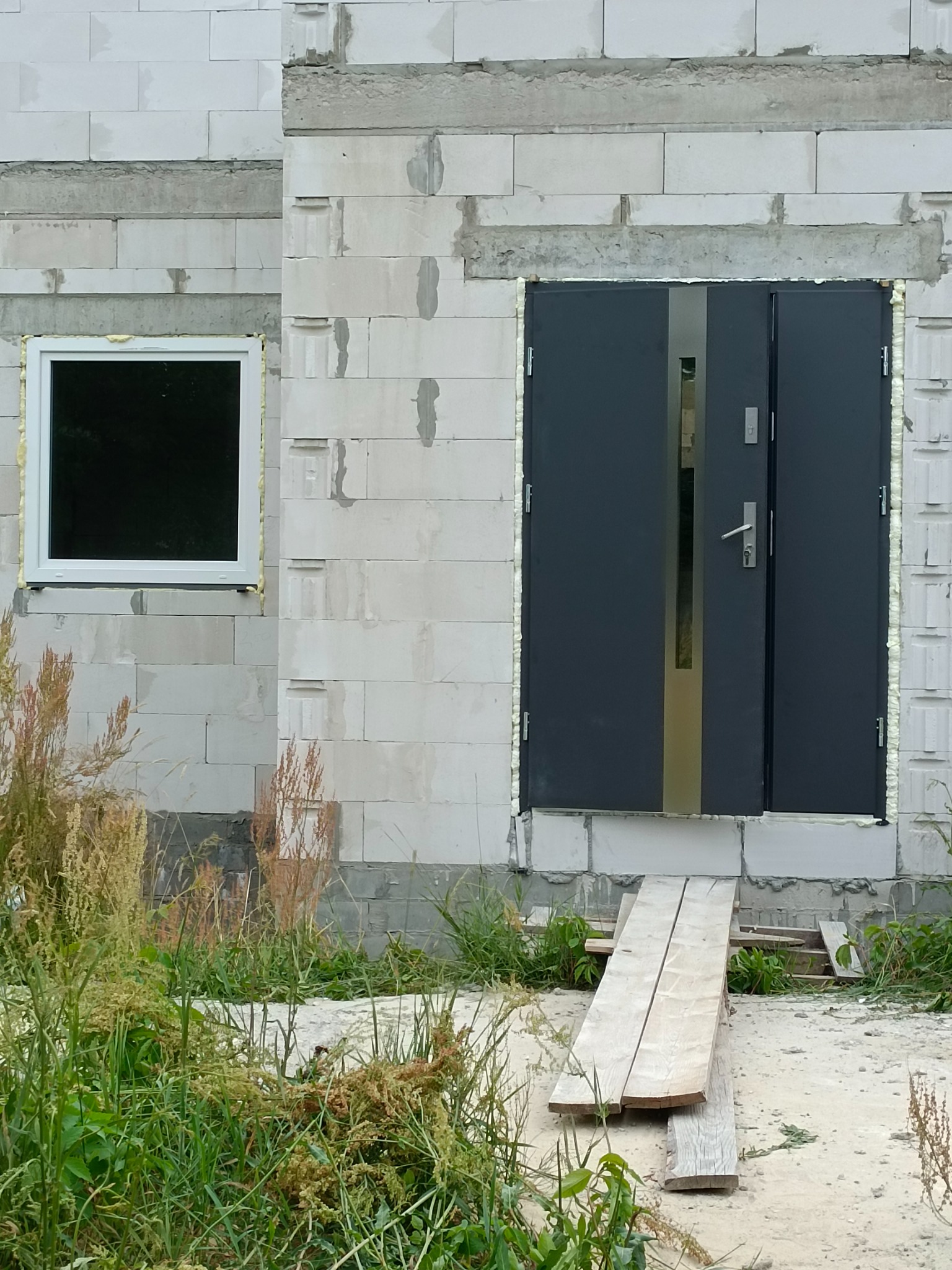 Nowe, grafitowe drzwi wejściowe z pionowym przeszkleniem i białe okno PCV w surowym stanie budynku z betonu komórkowego, widoczne pianowanie uszczelniające.