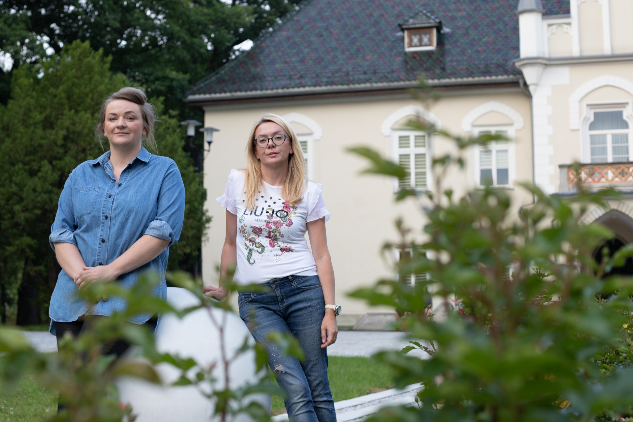 Dwie kobiety, blondynka w okularach oparta o biały element architektoniczny, druga w bluzie jeansowej, na tle budynku z wieżyczką i ogrodem.