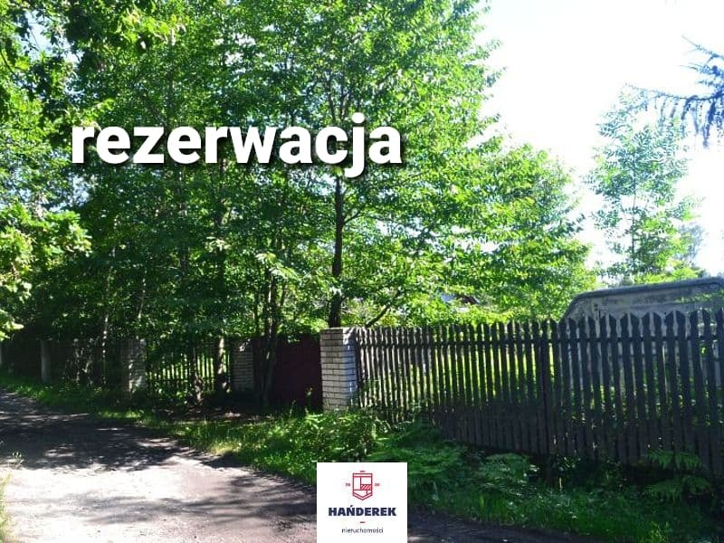 Działka z drewnianym ogrodzeniem i tabliczką 'rezerwacja' na tle zieleni, logo agencji nieruchomości w rogu.