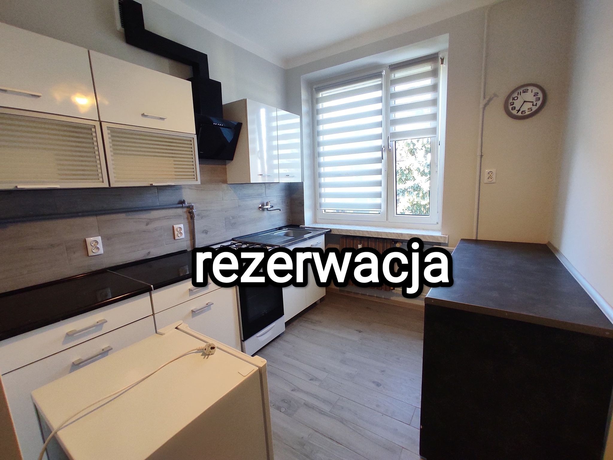 Jasna kuchnia z białymi szafkami, czarnym okapem i blatem, drewnopodobnymi płytkami na ścianie i podłodze, roletami w oknie i zegarem na ścianie. Na zdjęciu widoczny napis 'rezerwacja'.